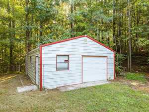 A small, white metal garage with red trim sits on a grassy lot, surrounded by tall trees. The building has a single white garage door, one window, and a posted sign visible in the front window.