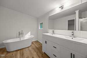 A modern bathroom with a freestanding white bathtub, wood-look flooring, a double-sink vanity with a large mirror, and white walls. A small window and a shower are also visible.