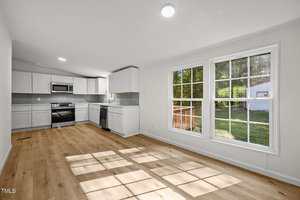 A modern kitchen with white cabinets, stainless steel appliances, and light wood flooring. Large windows on the right let in sunlight and offer a view of a grassy backyard with trees and a small outbuilding.