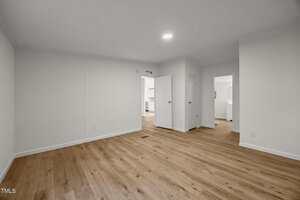 A spacious, empty room with light wood flooring, white walls, and a recessed ceiling light. Multiple doorways lead to other rooms, and there is visible trim along the walls and floor.