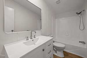 A modern bathroom with a white vanity, rectangular mirror, white toilet, and a bathtub-shower combination with a handheld showerhead. The floor is wood-toned, and the walls are white.