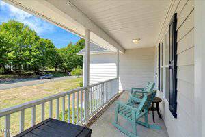 A covered front porch with two light blue rocking chairs, a small side table, and white railing, overlooking a grassy yard and a street lined with trees on a sunny day.