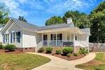Charming single-story white house at 2038 Ferbow Street in Creedmoor, featuring a gray shingled roof, brick foundation, and a covered front porch with white railings. Manicured shrubs line the curved walkway leading to the entrance.