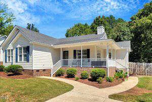 Charming single-story white house at 2038 Ferbow Street in Creedmoor, featuring a gray shingled roof, brick foundation, and a covered front porch with white railings. Manicured shrubs line the curved walkway leading to the entrance.
