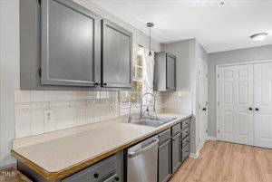 A kitchen with gray cabinets, a double sink under a window with a curtain, beige countertops, white tile backsplash, stainless steel dishwasher, and light wood flooring. Double doors are visible in the background.