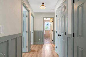 A hallway with light wood flooring, light blue paneled doors and wainscoting, off-white walls, and a ceiling light. At the end of the hall is a room with a window and white curtains that lets in natural light.