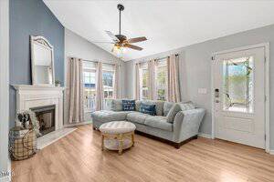 A living room with light gray walls, wood flooring, a sectional sofa, round ottoman, white fireplace, wall mirror, decorative pillows, and multiple windows with beige curtains letting in natural light.