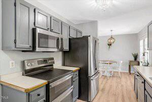 A kitchen with gray cabinets, stainless steel appliances, light wood flooring, and a view into a dining area with a round table, white chairs, and a modern chandelier.