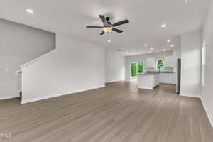 Spacious open-concept living area with light wood flooring, white walls, a ceiling fan, staircase, and a modern kitchen featuring white cabinets, an island, and sliding glass doors leading outside.