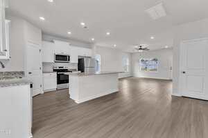 A modern kitchen with white cabinets, stainless steel appliances, and an island opens to a spacious living area with wood-style flooring and multiple windows. The room is well-lit with recessed lighting.