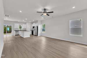 A spacious, unfurnished open-plan living area with light wood flooring, white walls, a ceiling fan, recessed lighting, and an adjacent kitchen featuring white cabinets and stainless steel appliances. Large windows provide natural light.