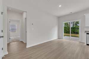 A room with light wood flooring, white walls, a sliding glass door leading outside, and an open doorway to another room with a window. The space has recessed lighting and a thermostat on the wall.