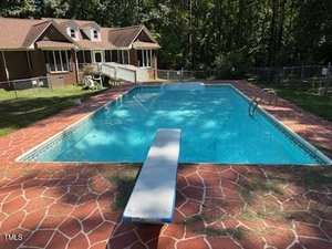 A rectangular outdoor swimming pool with a diving board is surrounded by a red stone-patterned deck at 80 White Deer Lane in Henderson. A brick house with a screened porch sits in the background, with a wooded area beyond the fenced yard.
