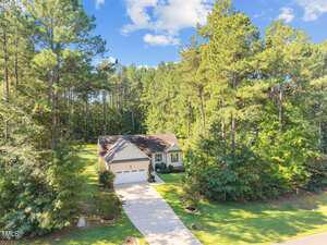 A single-story house with a two-car garage sits at the end of a long driveway, surrounded by tall pine trees and lush greenery under a partly cloudy sky.