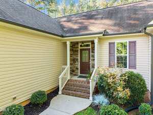 A beige house exterior with vinyl siding, brown shutters, and a covered front porch featuring brick steps, a dark brown door with glass panels, and landscaped bushes along the walkway.