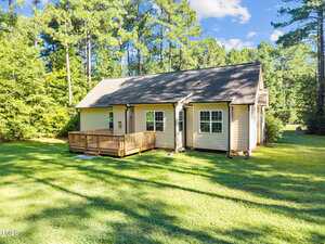 A small beige house with a gabled roof and a wooden deck in the backyard, surrounded by green grass and tall pine trees under a blue sky with scattered clouds.