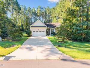 A single-story house with a two-car garage and a long driveway, surrounded by green grass, trees, and a wooded area in the background under a clear blue sky.