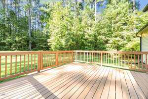 A wooden deck with railings and a view of a grassy yard bordered by tall green trees under a bright blue sky. Sunlight casts shadows on the deck floor.