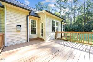 A wooden deck attached to a beige house with white trim, large windows, and a glass door. The deck is bordered by a wooden railing and faces a grassy yard with tall trees in the background.