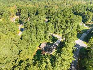 Aerial view of a suburban neighborhood with houses partially obscured by dense, mature trees, intersecting roads, and surrounding green forested areas.
