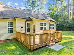 A yellow house with white trim features a wooden deck with railings and steps, set on a grassy lawn with trees in the background under a partly cloudy sky.