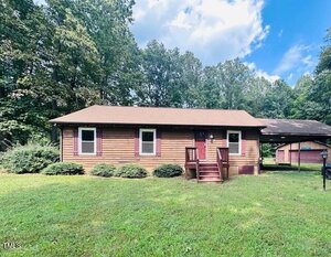 A single-story wooden house with brown siding and a front porch sits on a grassy lawn at 1545 Dogwood Drive, Oxford. Surrounded by trees, it features a small structure and covered area to the right. The sky is partly cloudy.