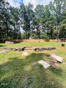 A grassy backyard with a small, circular stone fire pit in the center, a raised round pool to the left, a wooden fence in the background, and trees lining the perimeter. Some paving stones and wood are in the foreground.