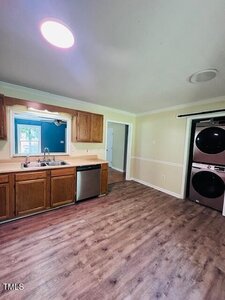 A kitchen with wood cabinets, a double sink, dishwasher, and wood-look flooring. In the adjacent area, a stacked washer and dryer is visible next to an open doorway leading to another room.