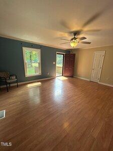 A room with wood flooring, a ceiling fan with light, blue accent wall with two windows, a maroon door open to a deck, a beige wall with a white door, and a chair in the corner. Sunlight streams through the windows.