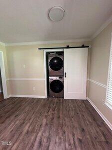 A stacked washer and dryer are in a closet with a white sliding barn door in a room with light beige walls, white trim, and wood-look flooring. A window is on the right wall.