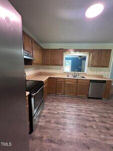 A kitchen with wood cabinets, a stainless steel refrigerator, oven, and dishwasher, laminate flooring, beige countertops, and a window above the sink overlooking an adjacent blue-walled room.