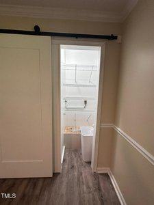 A small closet with white wire shelves and a white trash bin, viewed through a partially open sliding barn door. The walls are beige and the floor is dark wood.