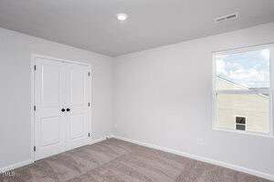 A small, empty room with beige carpet, white walls, a closed double-door closet, and a window letting in natural light. A ceiling light is on, and part of a building is visible outside the window.