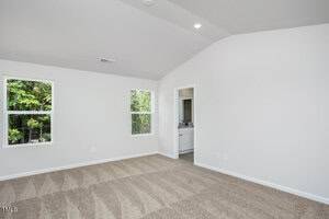 Empty room with beige carpet, white walls, two windows showing greenery outside, and a doorway leading to a bathroom with a double sink vanity. Ceiling is vaulted with recessed lighting.