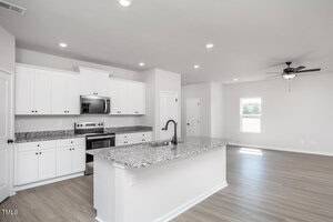 Modern kitchen with white cabinets, stainless steel appliances, granite countertops, a center island with a black faucet, and an open layout leading to a bright living area with wood-style flooring and a ceiling fan.