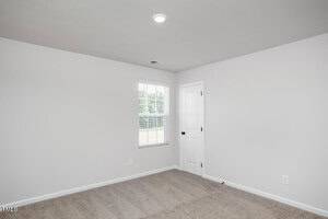 A small, empty room with light gray walls, a white ceiling, tan carpeting, a single window with blinds, and a closed white door. A ceiling light is turned on, and natural light enters through the window.