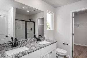 Modern bathroom with a granite countertop, double sinks, black fixtures, a large mirror, a glass-enclosed shower, a toilet, and an adjacent walk-in closet with shelves. A small window lets in natural light.