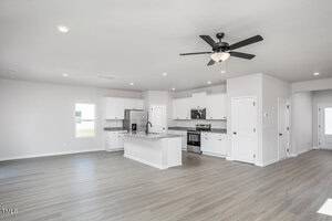 Open-concept kitchen and living area with light wood flooring, white cabinets, stainless steel appliances, a center island, and a black ceiling fan. Bright natural light comes in from a window.