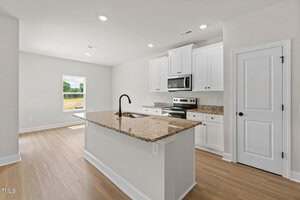 A modern kitchen with white cabinets, stainless steel appliances, a granite countertop island with a sink, and light wood flooring. A window in the background lets in natural light.