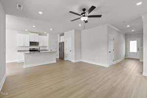 A spacious, empty kitchen and living area with light wood flooring, white walls, and ceiling. The kitchen features white cabinets, stainless steel appliances, and a granite countertop. A ceiling fan is mounted above the living space.