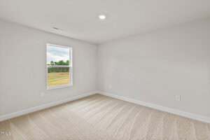 Empty room with beige carpet, off-white walls, a single window showing a grassy outdoor view, and a ceiling light. The room appears clean and well-lit with minimal decoration or furniture.