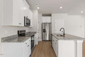 Modern kitchen with white cabinets, stainless steel appliances, granite countertops, an island with a black faucet, and light wood flooring under recessed lighting. Walls are painted a light beige color.