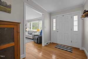 A bright entryway with a white front door, sidelights, and wood flooring. To the left is a wooden cabinet, and an open doorway leads to a living room with a couch, armchair, and large windows.