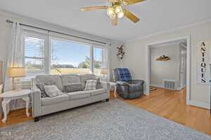 A living room with a beige sofa, plaid and patterned pillows, a gray recliner, side tables, a ceiling fan, large windows with blinds, hardwood floors, and a rug. A wall sign reads "ANTIQUES.
