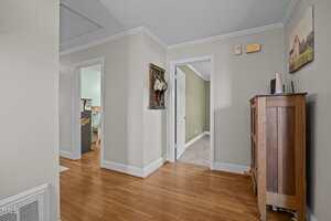 A hallway with light-colored walls, wood flooring, and white trim. Several doorways lead to other rooms, one painted green. A wooden cabinet and two framed pictures are visible along the walls.