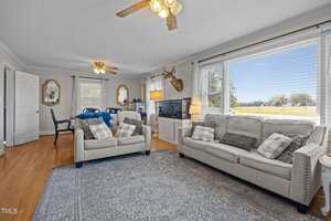A living room with two gray sofas, assorted pillows, a patterned rug, large windows with blinds, a ceiling fan, a mounted deer head, a TV on a stand, and a dining area in the background.