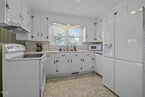 A kitchen with white cabinets, white appliances including a refrigerator, stove, and microwave, a coffee maker, a double sink under a window with blinds, and beige countertops and backsplash.