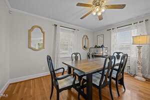 A dining room with a wooden table and six chairs, wood flooring, a ceiling fan with lights, two windows with curtains, a floor lamp, a mirror, and framed wall art. A small aquarium sits on a console table.