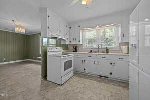 A kitchen with white cabinets, white appliances, and a double sink under a window with blinds. The floor is tiled, and an adjacent dining area with green walls and a chandelier is visible.