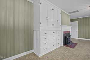 A room with green vertical paneled walls, white built-in cabinets with drawers and cupboards, a small black fireplace with a white mantel, and beige tile flooring. A closed white door is visible in the background.